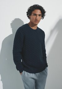 Mornarsko plavi džemper pleten s kablovima s dugim rukavima, okruglim izrezom i teksturiranim uzorkom, uparen sa svijetlosivim trenirkama.