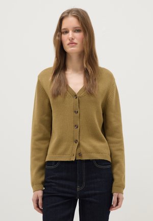Young woman standing wearing mustard buttoned cardigan and dark blue jeans against plain light background.