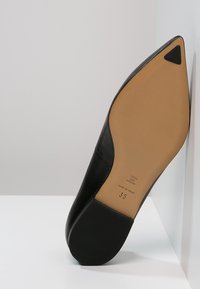 Zapato de tacón alto de cuero negro con puntera puntiaguda y superficie texturada. La suela es de color bronceado, mostrando información de tallas y la marca.