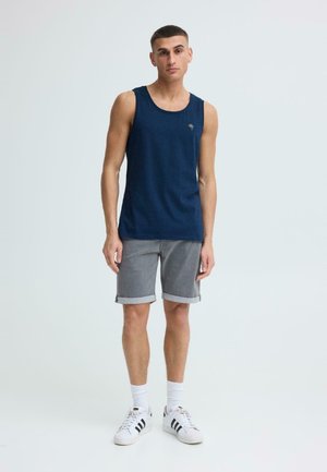 Joven de pie, vestido con una camiseta sin mangas azul marino, pantalones cortos grises remangados, calcetines blancos y zapatillas blancas con rayas negras sobre un fondo liso.