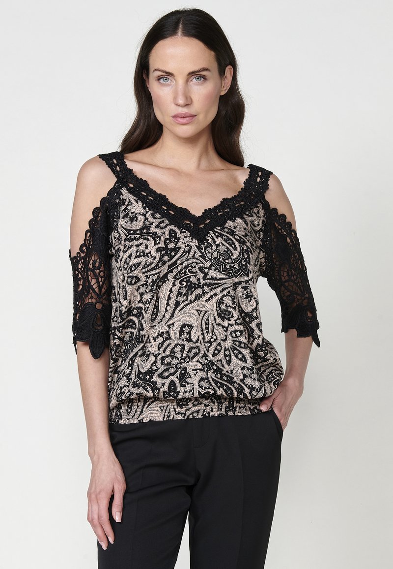 Top nero e beige con motivo paisley, maniche corte con spalle scoperte, caratterizzato da un intricato bordo in pizzo nero attorno al colletto e alle maniche.