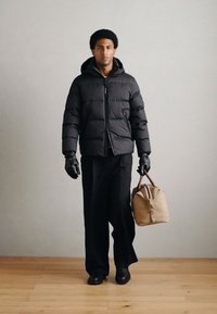 Doudoune noire avec fermeture éclair, associée à un pantalon noir et des gants. Tient un sac structuré marron clair avec deux poignées.