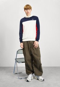 Felpa color block con sezioni blu navy, rosso e crema; cappuccio con stringhe; abbinata a pantaloni cargo larghi oliva; sneakers bianche.
