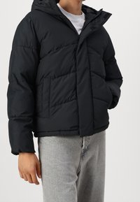 Black puffer jacket with a hood, featuring a zip closure, quilted texture, and two front pockets. Worn over a white shirt and gray pants.