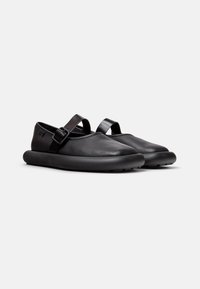 Zapatos de cuero negro con punta redonda, suela de goma plana y una única correa ajustable en la parte superior. Acabado suave con una marca sutil.