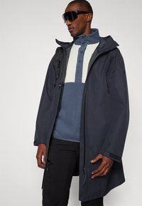 Manteau noir imperméable avec capuche, doté d'une fermeture éclair à l'avant, de poignets ajustables et d'un design léger. Porté par-dessus un pull bleu et blanc.