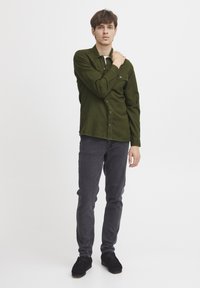 Zelena košulja s dugim rukavima i džepom na prsima, uparena s tamnosivim slim-fit trapericama i crnim cipelama, stoji ispred jedinstvene bijele pozadine.
