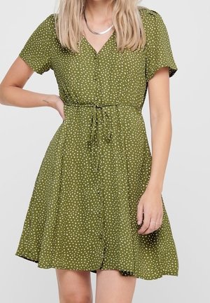 Femme portant une robe courte à manches courtes de couleur vert olive avec de petites taches blanches, une fermeture à boutons à l'avant et une ceinture nouée à la taille.