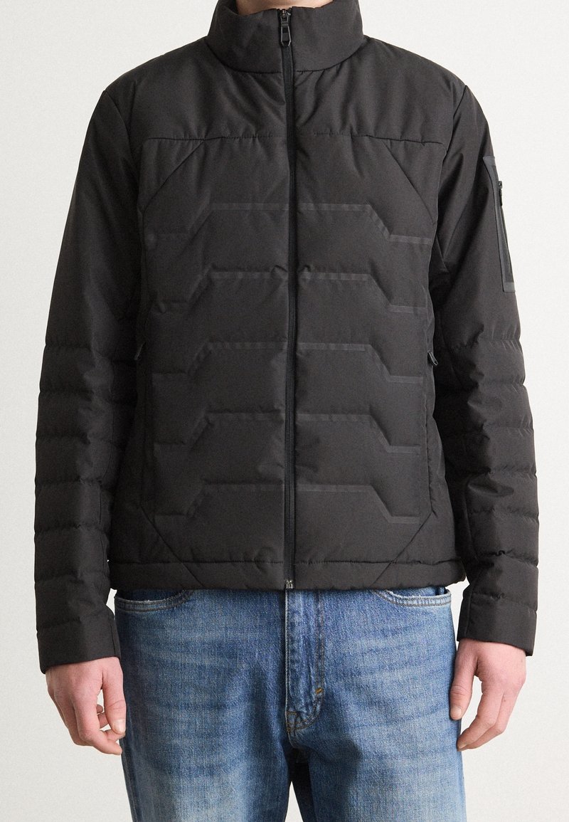 Mężczyzna w czarnej pikowanej kurtce puchowej z wysokim kołnierzem i zamkiem błyskawicznym, ubrany w niebieskie dżinsy, stojący na jednolitym tle.