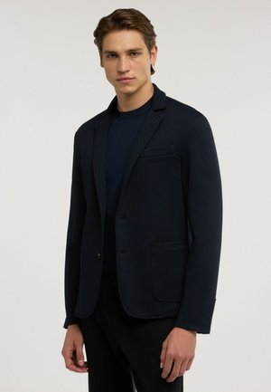 Ung man som bär en mörk marinblå blazer över en matchande skjorta, stående framför en enfärgad ljus bakgrund, tittar framåt med ett neutralt uttryck.