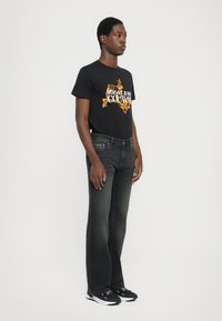 Črno grafično majico s kratkimi rokavi, ki nosi dizajn "VERSACE JEANS COUTURE", je kombinirana s temno sivimi jeans hlačami in črnimi tenisicami, stojijo na nevtralni ozadju.