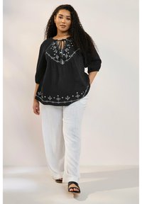 Svart, broderad blus med trekvartsärmar, knytkrage och mönstrad nederdel, ihopparad med vita, löst sittande byxor och svarta sandaler.