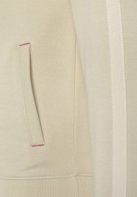 Šviesiai smėlinis džemperis, turintis tekstūruotą paviršių, šoninį kišenę su rožine siūle ir lygią baltą akcentinę juostą palei siūlę.