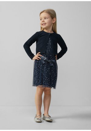 Navyblaues Kleid mit weißen Punkten, kurzer Tüllrock und einem passenden Schleifen-Gürtel. Kombiniert mit einem schwarzen Cardigan mit langen Ärmeln und silbernen Schuhen.