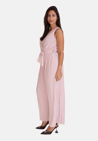 Lichtroze jumpsuit met een wikkelontwerp, geplooide textuur en tailleband met strikdetail. Gepaard met zwarte schoenen met spitse neuzen en strikaccenten.