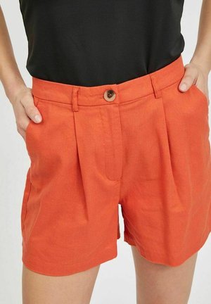 Kvinde iført orange shorts med høj talje, for-pliseringer og knaplukning, hænderne i lommerne, kombineret med en sort top.