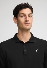 Svart polotröja i texturerad tyg, med en tre-knapps knappslå och en liten vit logotyp på vänster bröst.