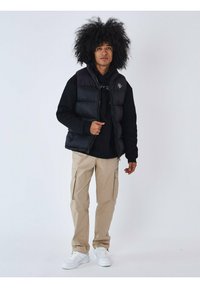 Chaleco de plumón negro sobre una sudadera negra, pantalones cargo beige y zapatillas blancas. Las características notables incluyen detalles de cremalleras y un logo en el chaleco.