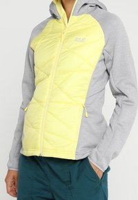 Lätt, vadderad jacka med gul framsida och grå ärmar. Har en huva, dragkedjefickor och elastiska muddar. Mjuk, andningsbar textil.