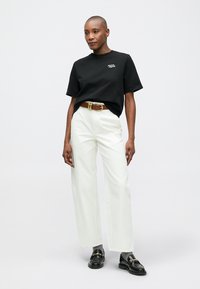 Czarny bawełniany t-shirt z małym białym logo, w parze z białymi spodniami o szerokich nogawkach i czarnymi loafersami, z brązowym paskiem.