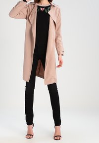 Trench coat nudo con apertura frontale, maniche lunghe e spalline. Indossato sopra un top nero con decorazioni e jeans skinny neri. Tacchi neri.