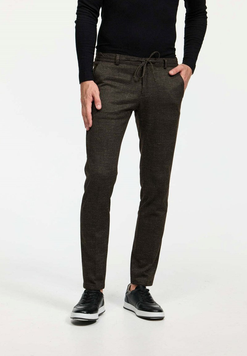 Braune, strukturierte Hose mit einem Kordelzugbund und schmal zulaufenden Beinen, kombiniert mit schwarzen Sneakers mit weißen Sohlenakzenten.