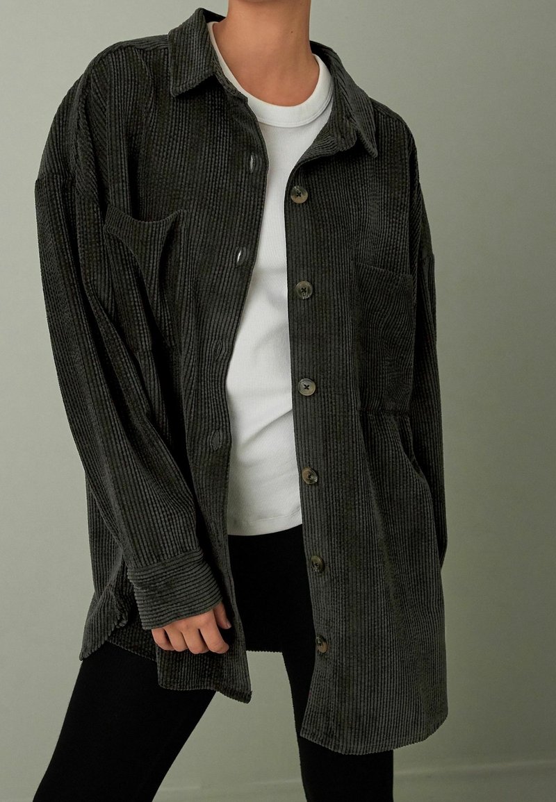 Personne portant une chemise surdimensionnée en velours côtelé gris foncé par-dessus une chemise blanche côtelée et un pantalon noir sur fond uni.