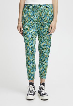 Floral patterned pants in green, blue, and yellow tones; elastic waistband with drawstring; tapered leg design; paired with black sneakers.