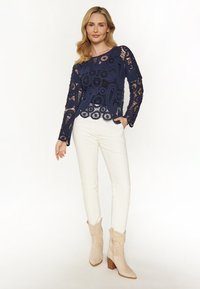 Top croșetat bleumarin cu mâneci lungi, modele complicate, tiv ondulat. Asortat cu pantaloni albi cu croi slim și cizme cu toc bej.