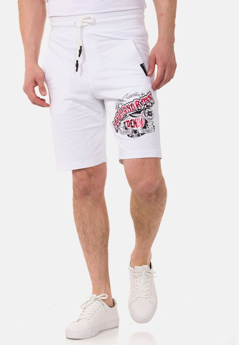 Weiße Shorts mit einem Kordelzug und Seitentaschen, mit rotem grafischen Druck auf dem linken Bein. Kombiniert mit weißen Turnschuhen.