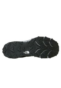Suela de zapato de goma negra con un dibujo grueso y texturizado, con tacos multidireccionales y un logo blanco de "The North Face."