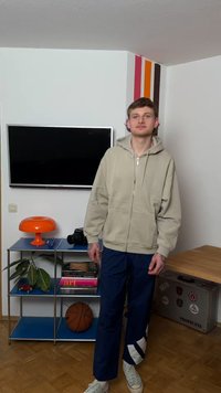 Ung mand står indendørs ved hylder med orange lampe, basketball, kamera og bøger, ved siden af et TV monteret på en hvid væg med farvede striber.