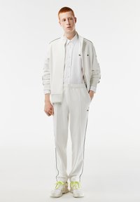 Survêtement de sport blanc avec des empiècements noirs, comprenant une veste zippée et un pantalon à taille élastique. Accompagné de baskets blanches et de lacets vert fluo.