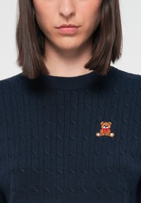 Granatowy sweter z splotem w warkocze z małym haftowanym misiem po lewej stronie klatki piersiowej. Ściągaczowy okrągły dekolt i miękka faktura.
