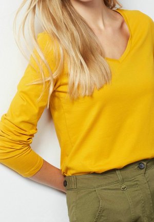 Femme blonde portant un shirt jaune à manches longues et col en V, rentré dans un pantalon taille haute vert olive avec des détails de boutons.