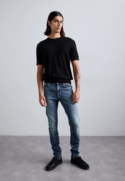 Schwarzes T-Shirt mit kurzen Ärmeln, hellblaue Jeans und schwarze Sneaker. Lange Haare, Tattoos, steht auf einem grauen Boden vor einem schlichten Hintergrund.