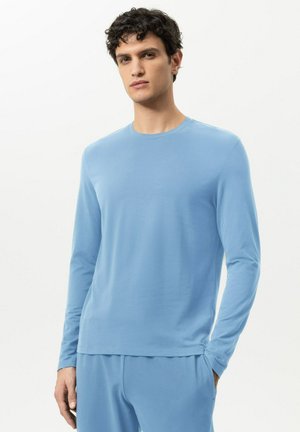 Mann mit dunkel lockigem Haar, trägt ein hellblaues Langarmhemd und passende Hose, eine Hand in der Tasche, vor weißem Hintergrund.