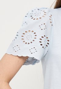 Λευκό κοντομάνικο με κυκλικό κέντημα με τρύπες και κυματιστό τελείωμα, που φοριέται από άτομο με ανοιχτόχρωμο δέρμα και μακριά καστανά μαλλιά.