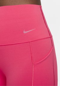 Różowe legginsy sportowe o wysokim stanie, wykonane z elastycznego materiału, z małym odblaskowym logo Nike i bocznymi kieszeniami do przechowywania.