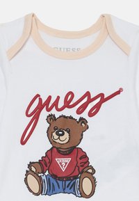 Tutina bianca in cotone con una grafica di un orso bruno che indossa un maglione rosso e jeans blu, con la scritta "guess" in lettere corsive rosse sopra.