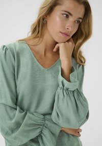 Blusa verde chiaro con scollatura a V e maniche a sbuffo arricciate. Realizzata in tessuto morbido con una texture liscia, presenta una vestibilità comoda.