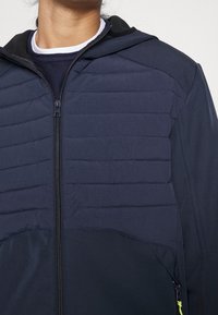 Marinblå jacka med ett vadderat panel fram, full dragkedja och huva. Har slätt tyg, sidofickor och kontrasterande dragkedjedetaljer.