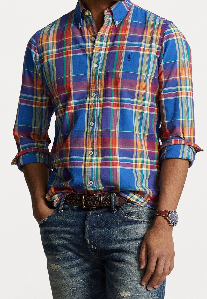Homme portant une chemise à carreaux multicolore boutonnée avec les manches retroussées, un jean bleu délavé, une ceinture tressée marron et une montre-bracelet.
