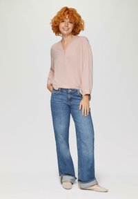 Lichtroze long-sleeve top met v-hals en borstzak, gecombineerd met blauwe wijde jeans. Het model draagt beige platte schoenen met een textuurpatroon.