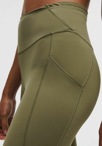 Detailní záběr osoby v olivově zelených legínách s vysokým pasem, které mají boční kapsu a viditelné švy.