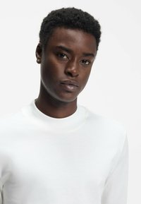 Weißes Langarmhemd mit hohem Kragen, aus glattem Stoff. Das Design zeichnet sich durch einen minimalistischen Stil mit einer taillierten Silhouette aus.