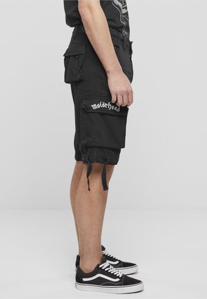 Isik, kes kannab musta cargo lühikesi pükse tekstiga „Motörhead“ taskul, musta graafilise mustriga T-särki ja must-valgeid tossusid, seisab küljele.