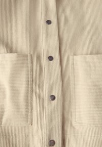 Beige korduroy bunda s vertikálním proužkovaným texture, čtyřmi předními knoflíky a dvěma velkými náprsními kapsami. Hladký povrch látky bez vzorů.