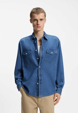 Jonge man met een open blauwe spijkerbroek overhemd over een wit ondershirt en beige broek, staand tegen een effen witte achtergrond.