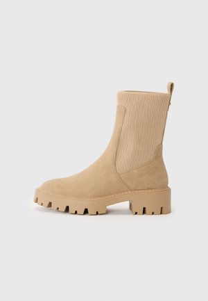Beige ankelstøvel i semsket materiale, ribbet elastiske sider, flat såle og kraftig hæl. Teksturert detalj forbedrer designet.